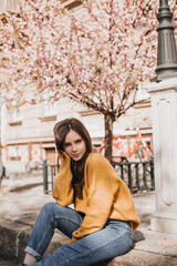 Woman in orange sweater sits on street road. Charming girl in jeans posing outside near bloomimg sakura. Lady looking into camera