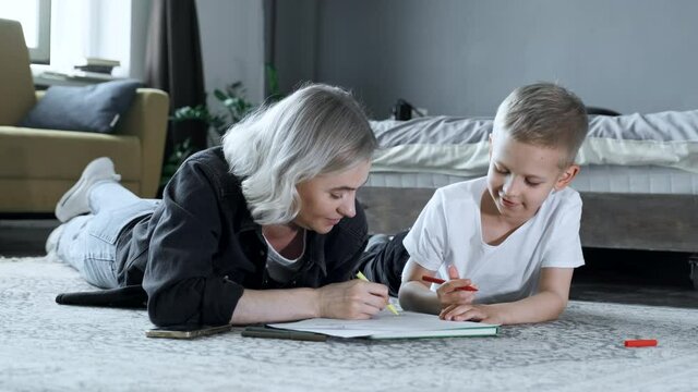 A Young Mother and A Baby Boy Are Sitting on the Floor, The Mother Is Teaching the Child To Draw, The Boy Is Drawing with Felt-Tip Pens on Paper, They Are Engaged in Training, Talking and Having Fun
