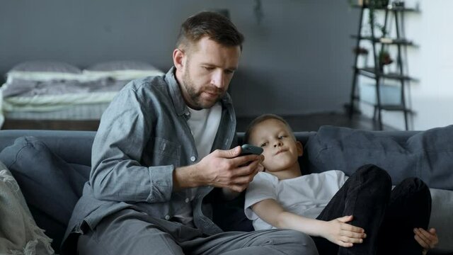 A Young Father and a Young Son, A Boy Child, Are Watching in the Smartphone Screen, Sitting on the Couch in a Cozy Room