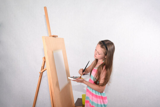 A beautiful girl artist of school age in the studio on a white background with a brush in her hand and watercolors paints a picture at an easel on paper