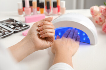 Woman using ultraviolet lamp to dry gel nail polish at white table, closeup - obrazy, fototapety, plakaty