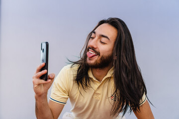 young hispanic latino male, long-haired, college student taking a selfie with his smart cell, smiling and sticking out his tongue © juanpablo