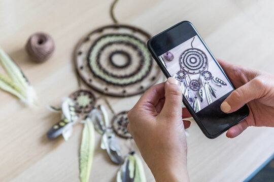 Woman craftsman photographing dreamcatcher use smartphone at workshop for internet advertising
