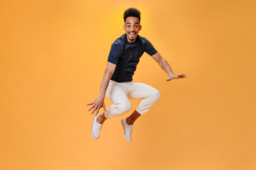 Fototapeta na wymiar Perky guy in white pants jumping on orange background. Portrait of brunette man in black tee posing on isolated backdrop