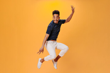 Fototapeta na wymiar Cheerful guy in black T-shirt jumping high on orange background. Snapshot of man in white pants dancing and having fun on isolated