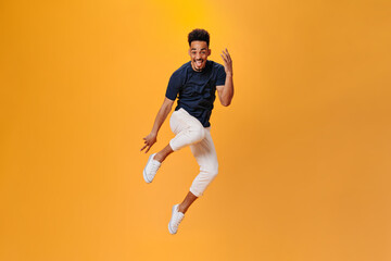 Obraz na płótnie Canvas Optimistic dark skinned guy in stylish outfit jumping on orange background. Brunette man in black t-shirt and white pants posing on isolated