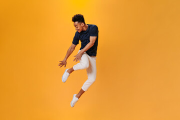 Fototapeta na wymiar Young man in white pants and black T-shirt jumping high on orange background. Brunette guy having fun on isolated backdrop