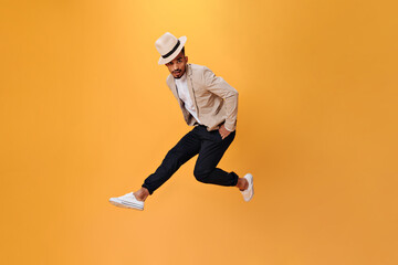 Fototapeta na wymiar Handsome guy in hat and suit is jumping high on isolated background. Young man in beige jacket and white shirt dancing on orange backdrop