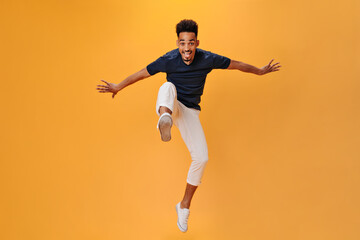 Fototapeta na wymiar Handsome man in black t-shirt smiles and jumps on orange background. Cool guy in white pants moving on isolated backdrop