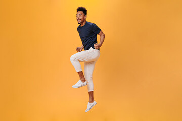 Young man in stylish outfit happily jumping on orange background. Portrait of guy in white pants moving on isolated backdrop