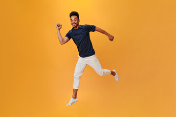 Fototapeta na wymiar Perky man in light pants jumping and having fun on orange background. Snapshot of brunette guy in white trousers posing on isolated