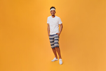Fototapeta na wymiar Man in sport outfit is smiling on orange background. Full-lenght portrait of guy in white tee and shorts posing on isolated backdrop