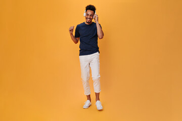 Fototapeta premium Stylish man in white pants listens to music in massive headphones. Portrait of guy in dark-blue t-shirt dancing on orange background