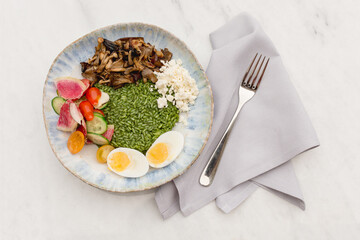 Vegetarian Green rice bowl, kale-almond pesto, oyster and maitake mushrooms, radishes, cherry tomatoes, feta cheese, and soft boiled eggs, on a white marble table with a fork on a gray napkin