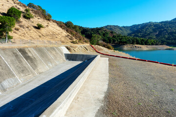 Concrete dam spillway, orange debris boom on dry ground. Extremely low water level in drying during summer Stevens Creek reservoir in San Francisco Bay Area, California