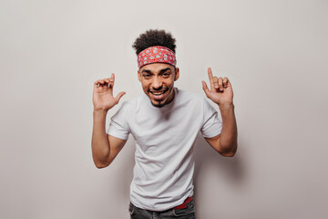 Fototapeta premium Cool guy in bandana looks into camera on white background. Portrait of man in white t-shirt and black jeans smiling and dancing on isolated