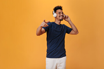 Young man in white headphones enjoys music on orange background. Cheerful guy in pants and blue tee listening to songs in headphones on isolated backdrop