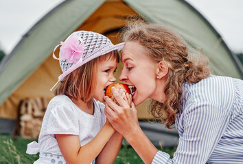 Mother and daughter eating apple near a tent in meadow or park. Happy family on picnic at camping