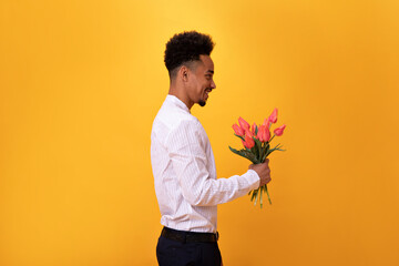 Charming man smiles and holds bouquet of flowers on orange background. Guy in white shirt is ready for romantic date and posing with tulips on isolated