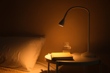 Glowing lamp and book on table in dark bedroom - obrazy, fototapety, plakaty