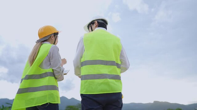 Low angle shot of two employees in high visible working gears discussing on working project, with background of natural mountain range and clear sky behind, a construction builder team, eco friendly 