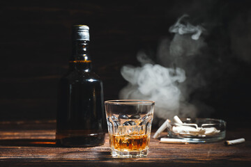 Bottle and glass of cold whiskey, cigarettes on wooden table against dark background