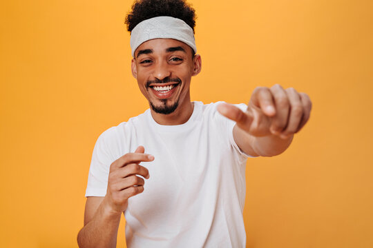 Stylish man in white T-shirt laughs and points his finger at camera. Portrait of brunette guy in white headband smiling and dancing on orange background