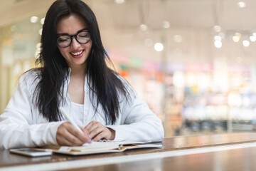 Happy Asian young woman sitting at cafe with coffee cup taking notes enjoying break