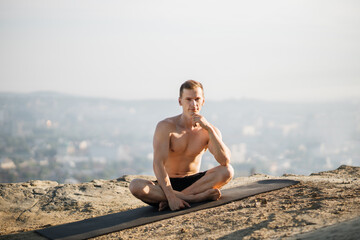 Fototapeta na wymiar Smiling joyful handsome caucasian man with bare torso sitting in lotus position on yoga mat and looking at camera. Healthy young guy meditating alone on high hill.