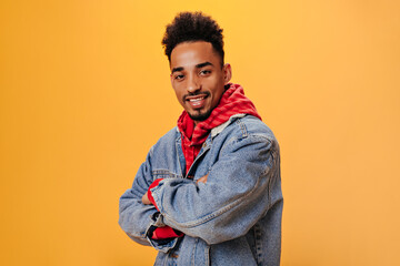 Stylish man in denim outfit posing on orange background. Curly brunette guy in red hoodie looking into camera on isolated backdrop