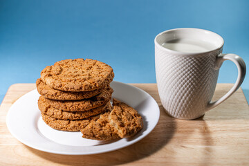 A mug with milk and oatmeal cookies on a wooden table. White plate with fresh oatmeal cookies on a blue background. Close-up.