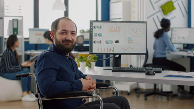 Sad businessman frustrated of his disabilities looking at camera sitting immobilized paralysed in wheelchair in business office working on pc with data, prosessing financial information