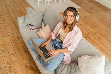 Young pleased happy  beautiful  woman sit indoors on sofa  using laptop computer listening music with earphones in living room . Top view.