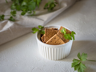 Natural flat rye mini-breads of a square shape in a white bowl on a light concrete background. Textile and parsley in the background. Delicious organic and quick snacks for diet meals