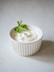 Close-up of fresh sour cream in white sauce bowl. Whirlpool of sour cream. Parsley leaves for decoration. Light concrete background. Delicious and natural dairy product