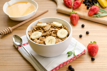 Healthy breakfast with bowl of cereals (muesli), strawberry, wild berries, honey and banana on light wooden table.