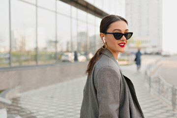 Stylish pretty woman with dark hair and red lips wearing sunglasses and jacket walking on city square in summer warm day
