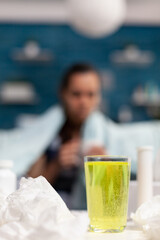 Young woman with illness drinking cup of vitamins as medical treatment for seasonal virus symptoms. Sick adult sitting on sofa at home wrapped in blanket with fever temperature