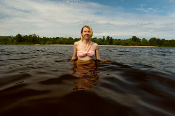 Fototapeta premium Portrait of wet blonde woman in swimsuit in dark water on nature. Swimming on the river