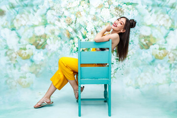 Obraz na płótnie Canvas Beautiful female mixed race model with pale skin, in a floral studio setting, wearing a yellow jumpsuit and using a chair to pose. 