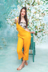 Naklejka premium Beautiful female mixed race model with pale skin, in a floral studio setting, wearing a yellow jumpsuit and using a chair to pose. 