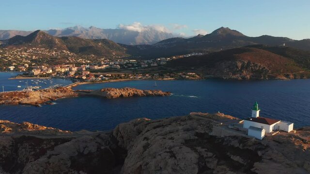 Aerial view of a desolated lighthouse in the middle of a desert island in the ocean, sea. Rocky shore and blue transparent water during sunset, mountain range and small city in the background, clouds