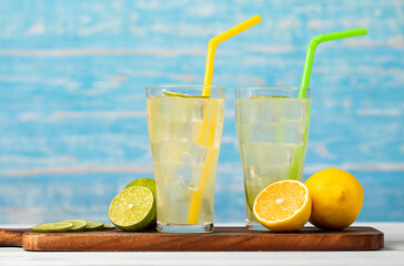 Refreshing glasses of cold fruit juice, ice lime juice, decorated using  fresh lime and lemon slice with yellow and green straw on wooden chopping board on white table in front of blue background
