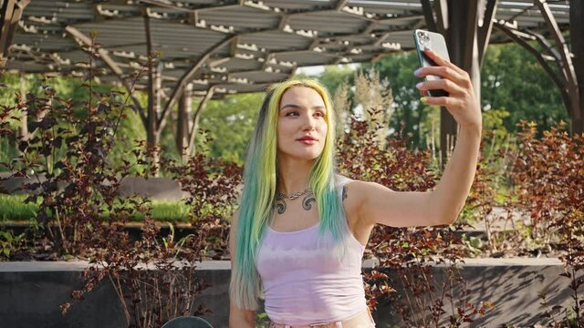 Young beautiful girl makes a selfie in the city park. Female teenager with tattoos, piercings and bright hair taking pictures of the outside