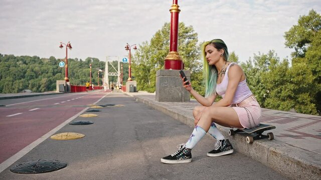 Female teen skateboarder with a bright appearance, tattoo and colored hair sits in a city park near the bridge. A girl holding a smartphone in her hands