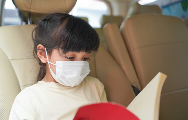 Asian little child girl wearing medical protective mask reading the book while sitting on the car during Covid-19 spreading. 