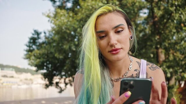 Close outside portrait of a young girl with a smartphone in her hands. A teenager with multi-colored yellow-blue hair, tattoos, piercings and a tunnel in the earlobe. Youth subculture concept