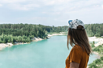 Rear view of a young blond woman traveler in a cap and mustard t-shirt standing on the cliff of blue turquoise lake or river and coniferous forest, travel nature beautiful landscape copy space tourism