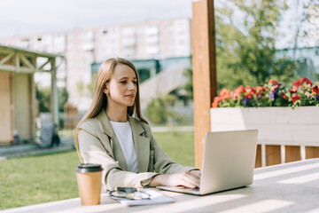 Beautiful blonde business woman working at laptop sit down on bench outside on a urban city street. Happy lady girl in suit distance learning, education and online shopping.