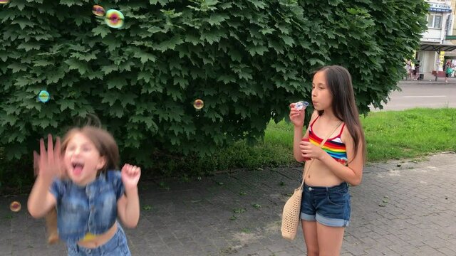 Two happy girls, sisters of ten and six years old, blow bubbles on the street in summer. 